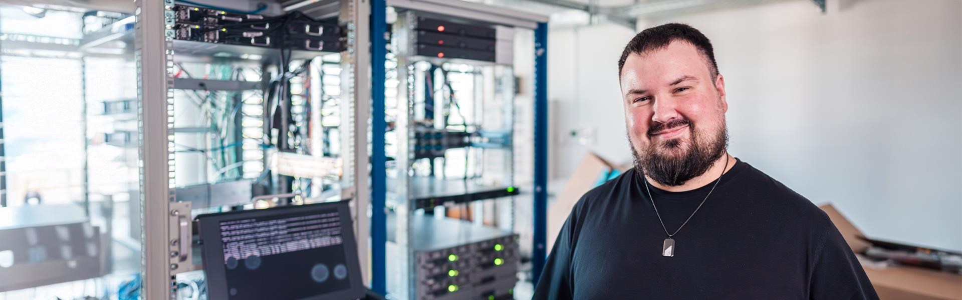 Person mit Bart steht in einem Serverraum und lächelt, im Hintergrund Serverracks und ein Monitor mit Codeanzeige.
