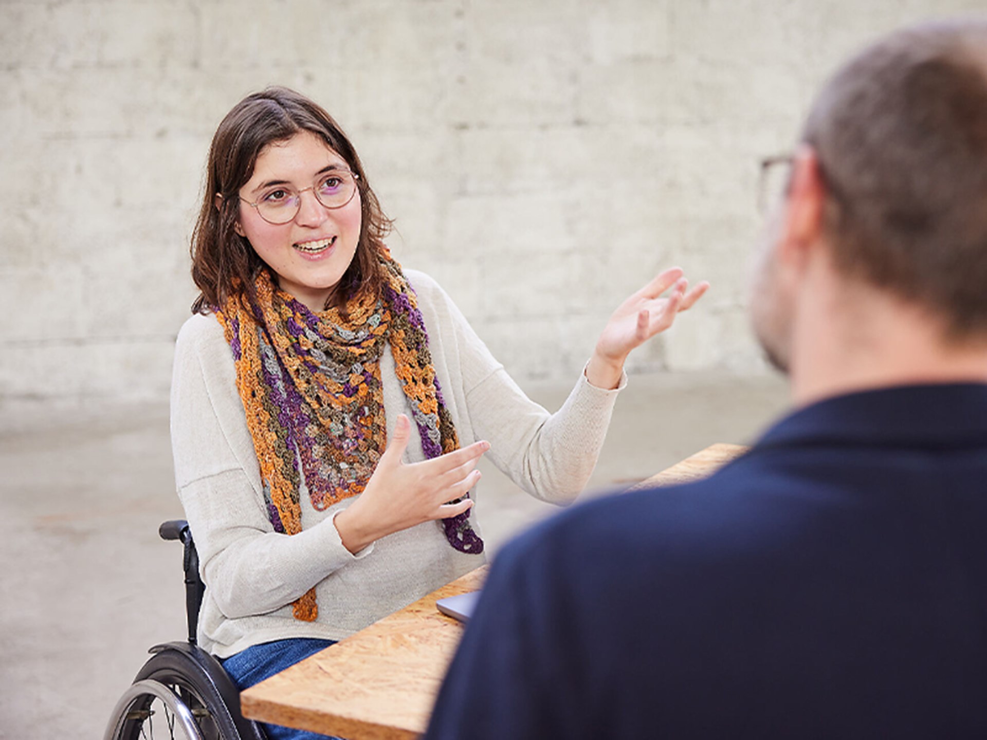 Rollstuhlnutzende Person spricht gestikulierend mit einer sitzenden Person in einem hellen Raum.